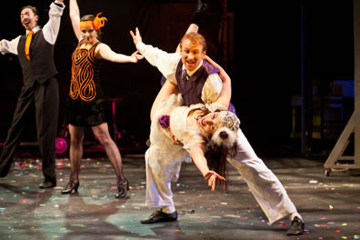 Orsino holding a flying Viola on his thigh, he in purple vest, with a dancing couple in the background, he in orange tie, and she in a black dress with orange scroll on the front and orange headpiece
